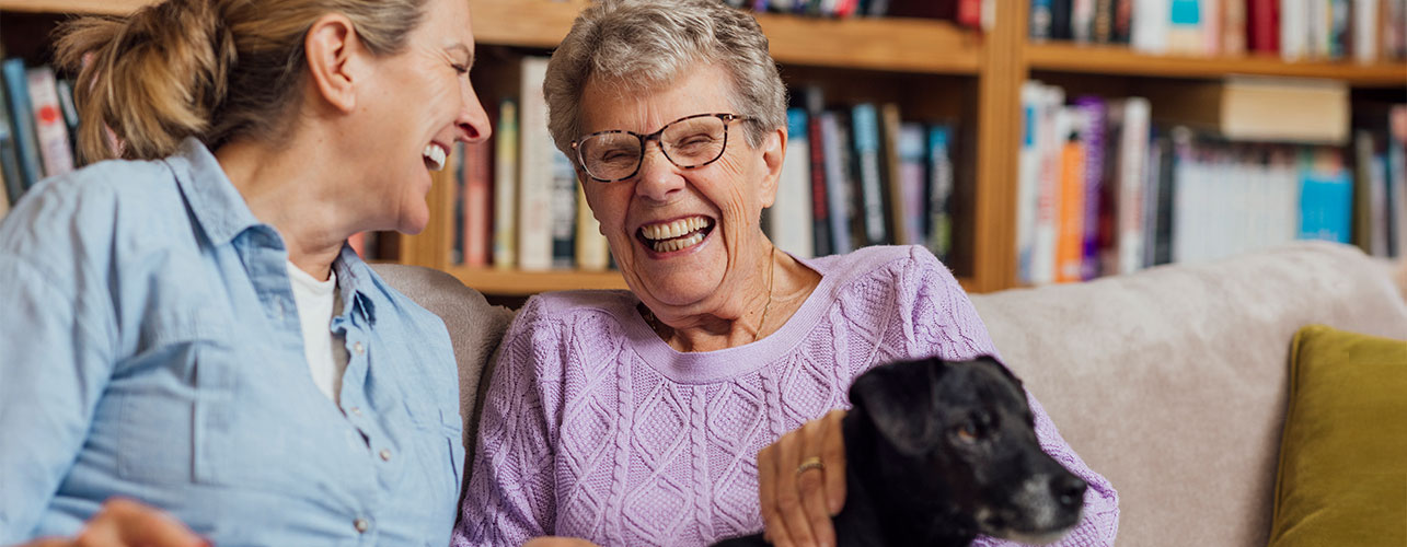 Mother and daughter sitting on a couch in a library with their small dog.